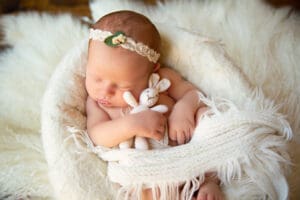 An infant baby sleeping with a bunny stuffed animal; swaddled in white linens and a white floral headband