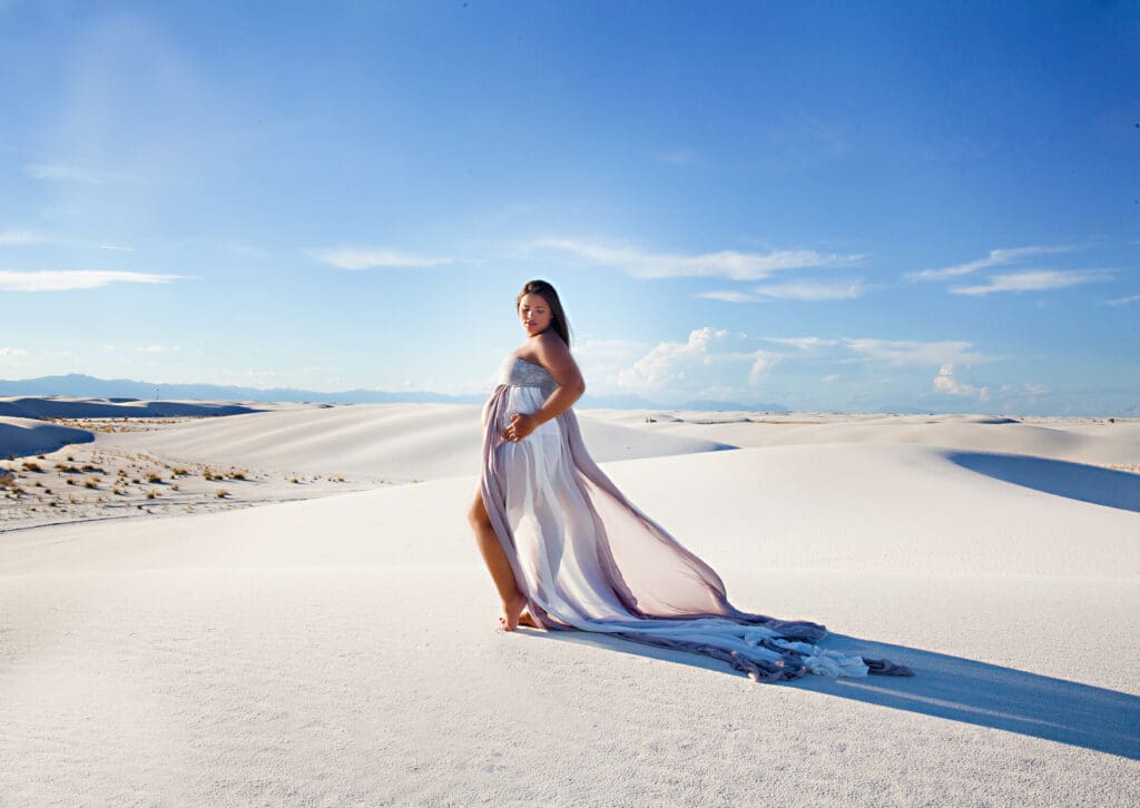 A woman in a flowing white dress standing in the sand on a clear blue sky