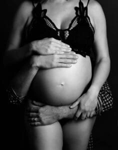 Black and white photo of a pregnant woman in a black, lace bra showing her baby bump with man's arms around her belly and her hands on top of the man's arms, photoshoot in Frisco Texas.